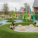 Children are playing in a modern, colorful playground area featuring rolling green mounds covered in artificial turf. In the foreground, three children are seated or kneeling around a sand-filled area with several light-colored, dome-shaped structures resembling buried eggs or hills. Tall, brightly colored vertical posts, some with a rainbow gradient (referred to as "gun barrel piling"), are scattered across the landscape. In the background, there are metal arches, a swing set, and a small wooden structure with a metal roof next to a canopy tent decorated with a large American flag star pattern